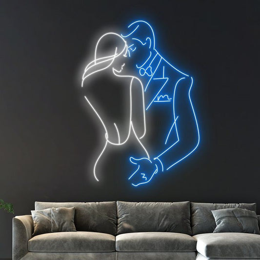 Romantic Couple Neon Sign – Custom Neon Sign for Living Room Neon light art of a couple embracing on a dark wall above a gray sofa.