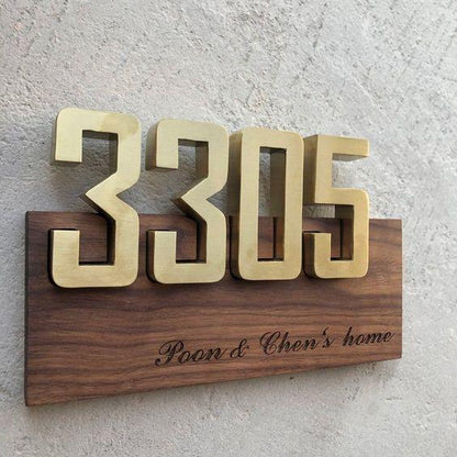 Luxury gold raised house numbers on a wooden address plaque with custom family text, displayed on modern grey stone and black marble exterior walls.