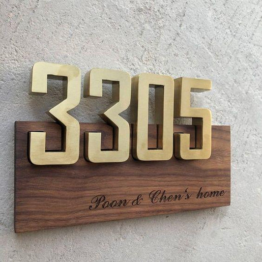 Luxury gold raised house numbers on a wooden address plaque with custom family text, displayed on modern grey stone and black marble exterior walls.