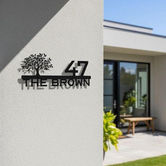 Premium personalised black metal family name sign with decorative tree silhouette, number 47, and Brown surname mounted on a contemporary stone entrance wall.