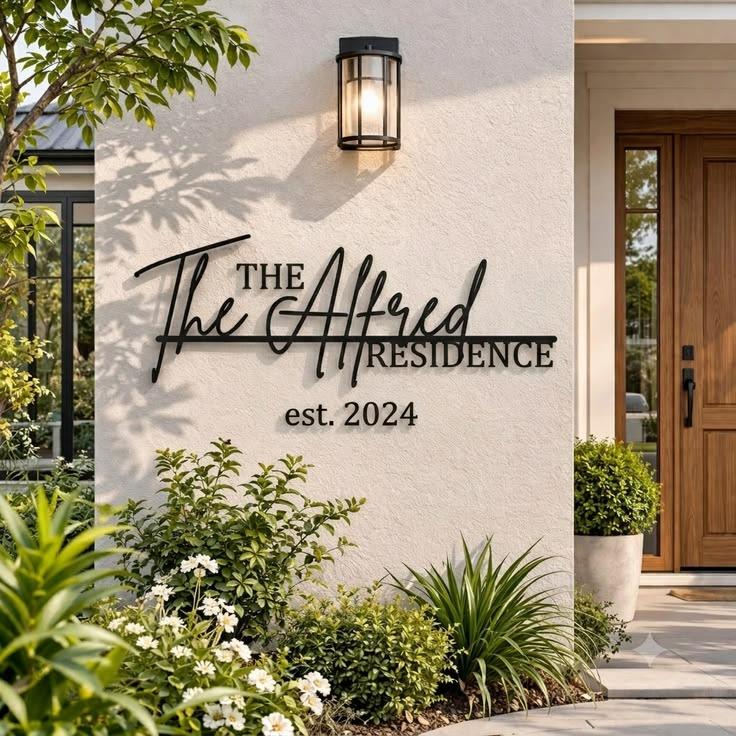 Modern personalised house name sign with raised black metal lettering and establishment year, mounted on wood cladding and textured stone entrance walls.