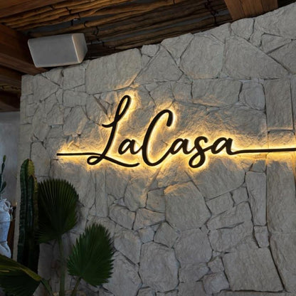 Modern illuminated house name sign with personalised cursive lettering and warm LED halo lighting mounted on a wooden wall and contemporary outdoor entrance.