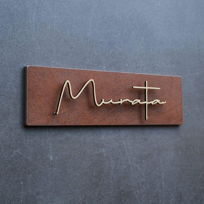 Modern personalised metal name sign with elegant gold script and a rustic brown finish displayed on dark stone and wooden entrance walls.