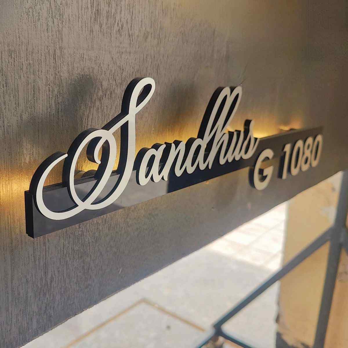 Modern illuminated family name sign featuring 3D cursive lettering and house number G 1080 against a premium beige stone wall.