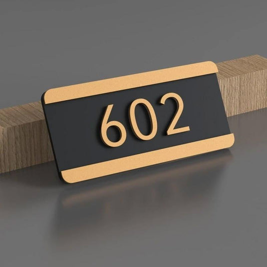 Modern rectangular black apartment number sign with gold 602 lettering and brushed gold borders mounted on a wood or stone entrance wall.