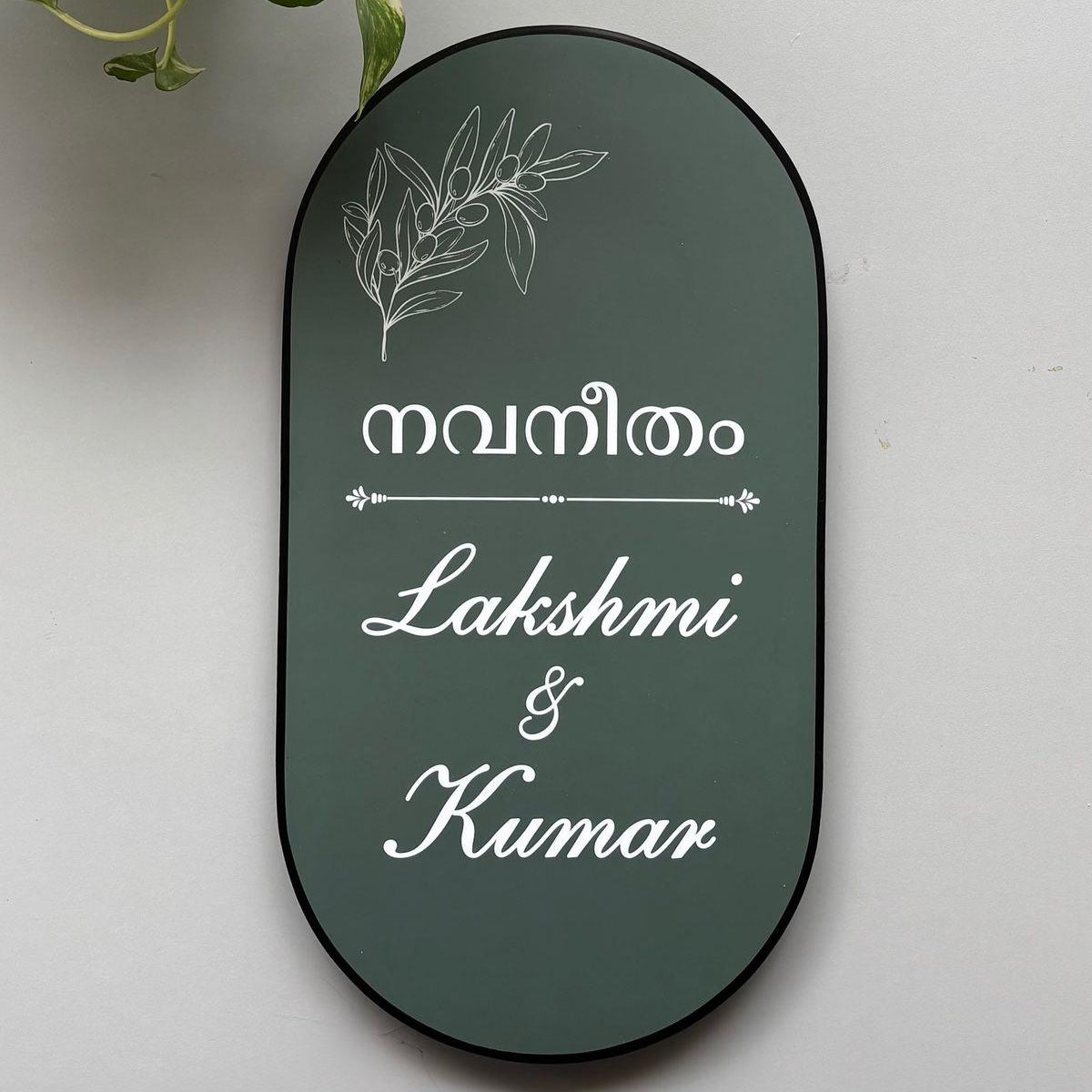 Premium personalised oval home name board with Malayalam text, botanical line art, white cursive lettering, and a black border mounted on a modern wooden entrance.