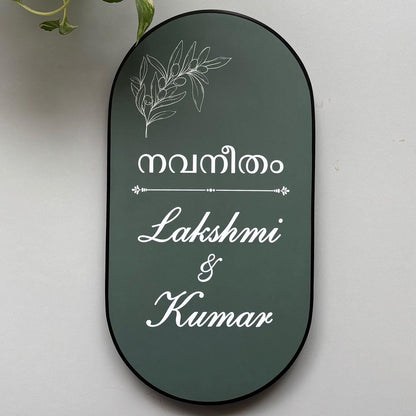 Premium personalised oval home name board with Malayalam text, botanical line art, white cursive lettering, and a black border mounted on a modern wooden entrance.