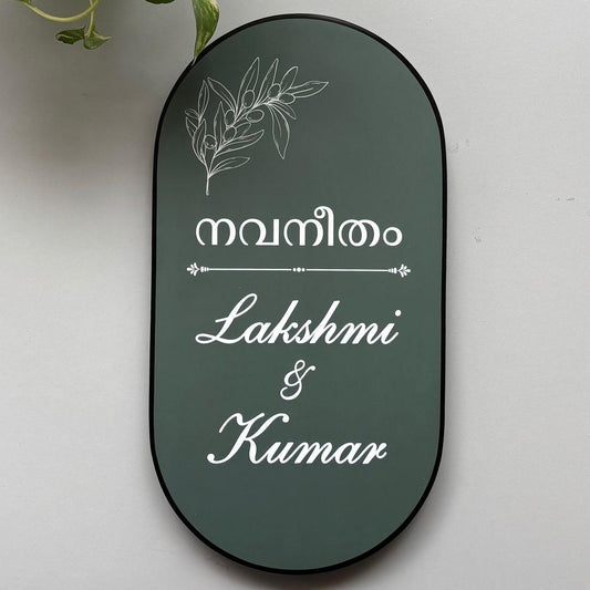 Premium personalised oval home name board with Malayalam text, botanical line art, white cursive lettering, and a black border mounted on a modern wooden entrance.
