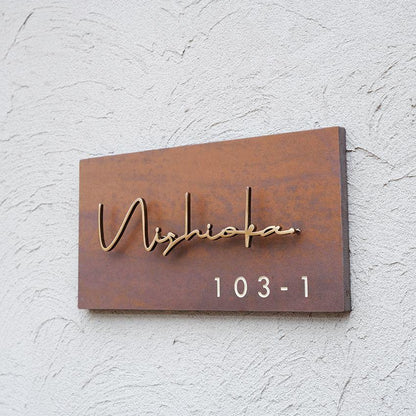 Modern rectangular brown metal house name sign with elegant gold cursive lettering and personalised address number, mounted on a contemporary stone entrance wall.