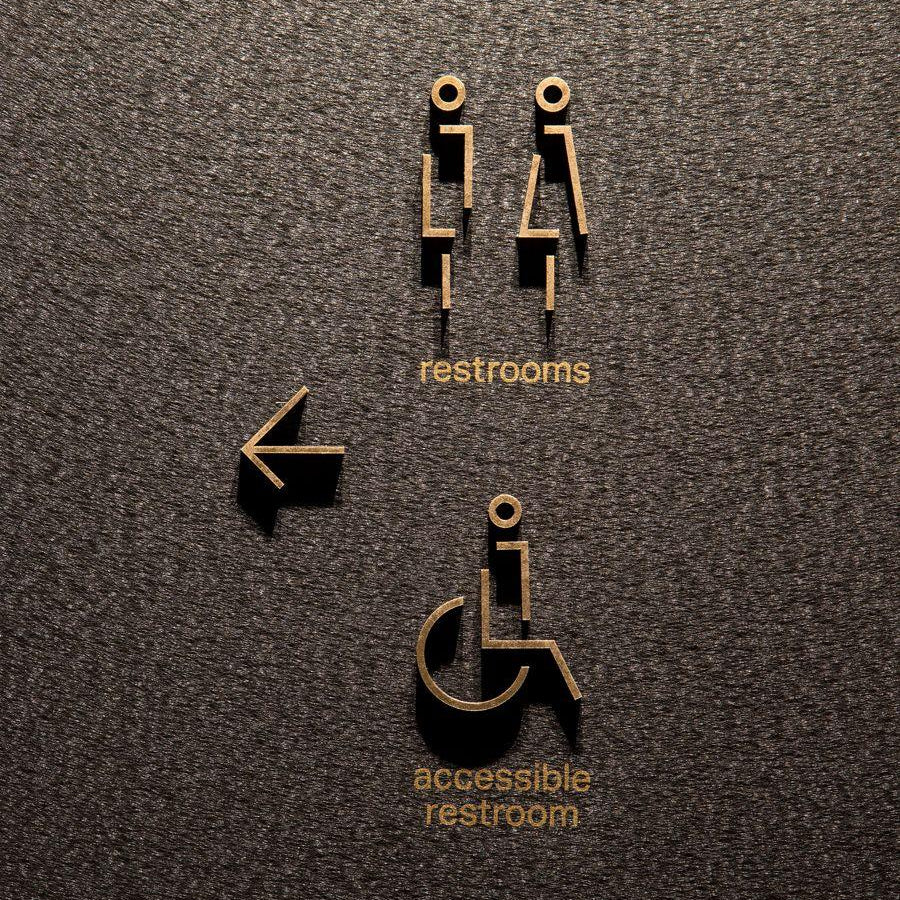 Modern gold restroom wayfinding signage with raised male and female symbols, accessible restroom text and directional arrow displayed on wooden, grey stone and marble walls.