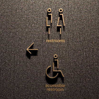 Modern gold restroom wayfinding signage with raised male and female symbols, accessible restroom text and directional arrow displayed on wooden, grey stone and marble walls.