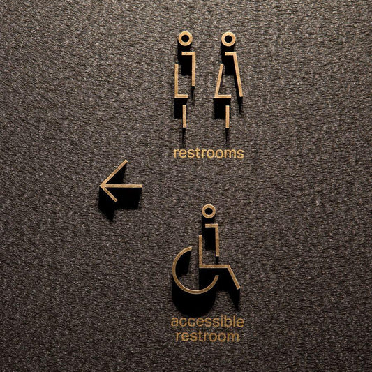 Modern gold restroom wayfinding signage with raised male and female symbols, accessible restroom text and directional arrow displayed on wooden, grey stone and marble walls.