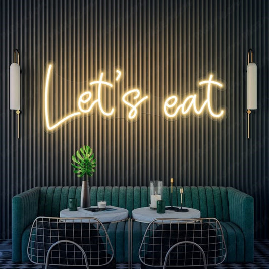 Lets Eat Neon Sign | Custom Neon Sign | Neon for Restaurant Neon sign reading 'Let's eat' above a modern dining area with green couch and decorative elements.