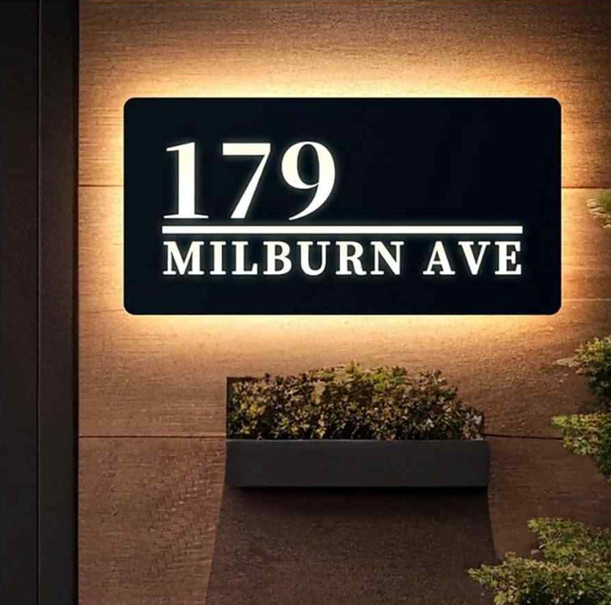 Illuminated black house number sign with white address lettering and warm LED backlight displayed on stone walls and wooden exterior cladding.