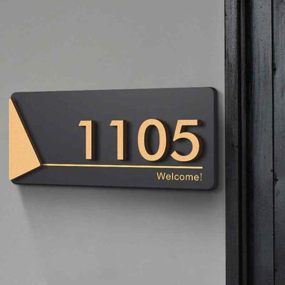 Premium black address plaque with gold 3D house numbers, triangular gold accent and personalized welcome message displayed on textured stone and wooden entrance walls.