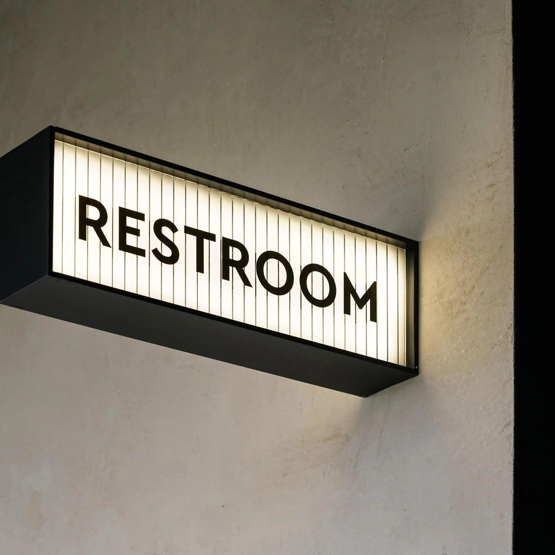 LED restroom signage with glowing white panel and black frame displayed on marble, wooden and textured walls in a contemporary commercial interior.