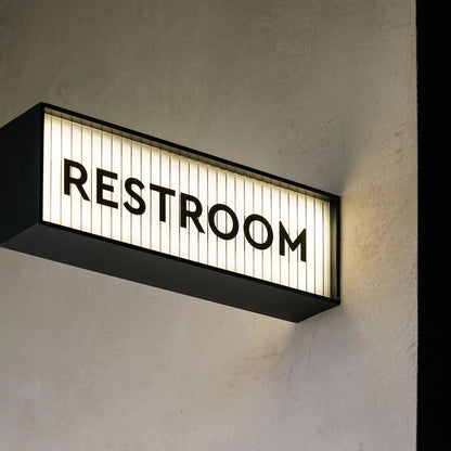 LED restroom signage with glowing white panel and black frame displayed on marble, wooden and textured walls in a contemporary commercial interior.