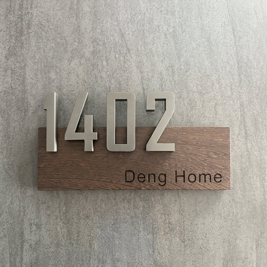 Custom 3D house number plaque with large dimensional address numbers and a personalised home name on a rectangular wood-effect sign displayed on exterior and timber-panelled walls.