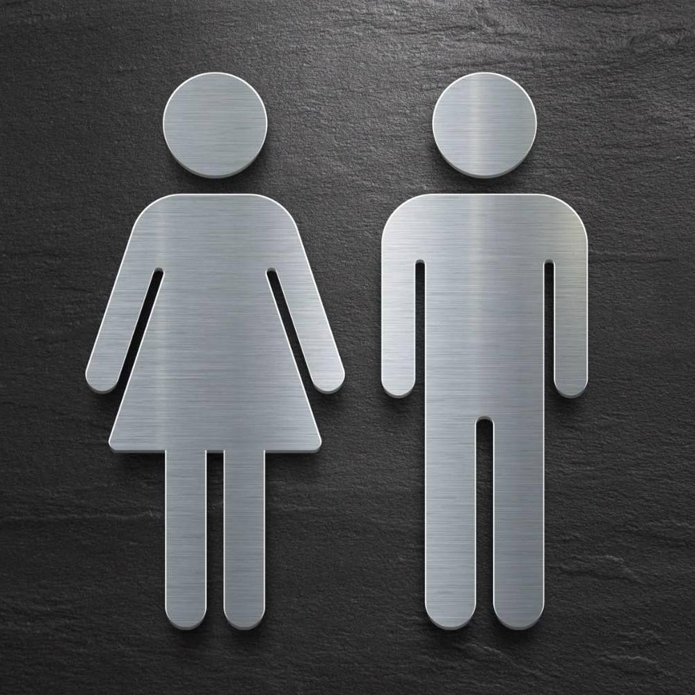 Brushed silver 3D-style toilet symbols for men and women displayed on beige marble, dark stone and grey textured walls in modern commercial interiors.