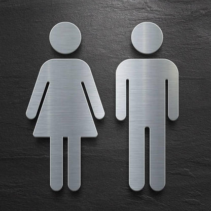Brushed silver 3D-style toilet symbols for men and women displayed on beige marble, dark stone and grey textured walls in modern commercial interiors.