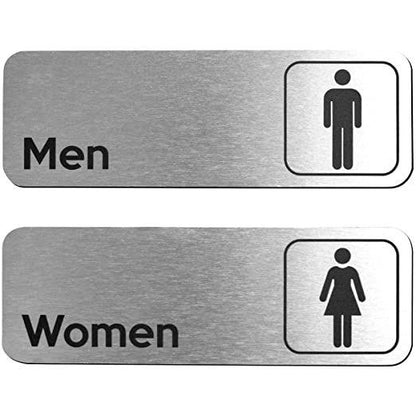 Brushed silver women's toilet sign with black text and female symbol displayed on beige stone, dark marble and wooden wall surfaces.