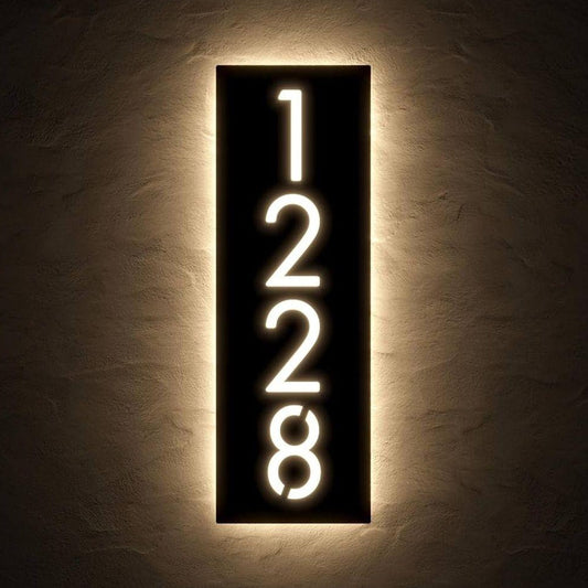 Premium black vertical address number plate with glowing white LED numerals and warm halo illumination, displayed on charcoal stone and wooden entrance walls.
