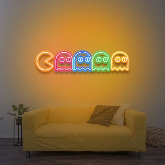 Pac Man Neon Sign  – Custom Neon Sign for Kids Living Room 30 By 15 Inches Neon Pac-Man ghosts sign on a wall above a yellow couch.
