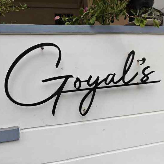 Custom script surname sign in black with flowing handwritten-style lettering, mounted on a neutral entrance wall with contemporary landscaping and greenery.