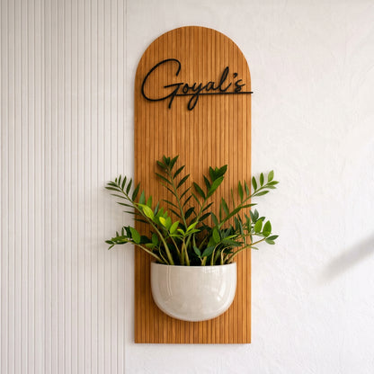 Modern decorative wall planter featuring a rounded wooden backdrop, custom black name lettering and a white plant pot for a contemporary home entrance.