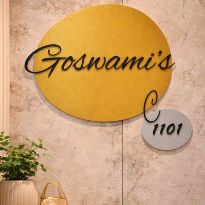 Modern circular gold nameplate with custom black cursive family name and matching apartment number plaque mounted on a stone and wooden entrance wall.