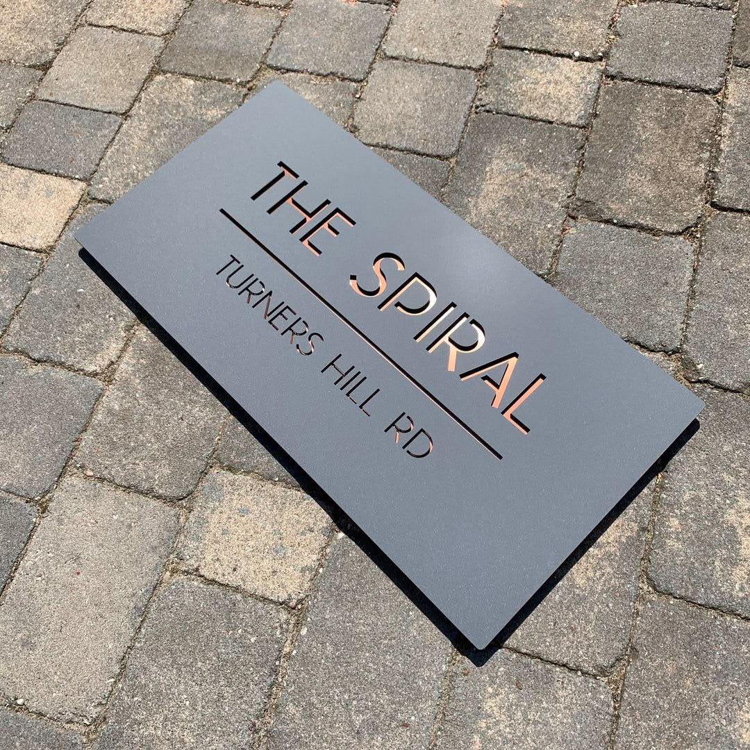 contemporary rectangular black house sign with metallic copper lettering and minimalist street address details mounted on wooden and marble-style walls.