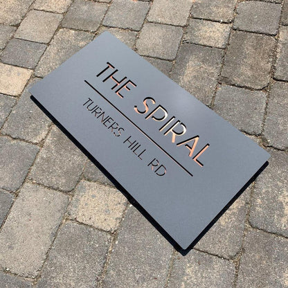 contemporary rectangular black house sign with metallic copper lettering and minimalist street address details mounted on wooden and marble-style walls.