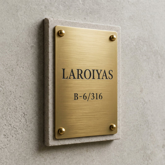 Modern rectangular gold address plaque displaying a customised family name and apartment number, mounted on a textured stone panel beside a wooden entrance door.