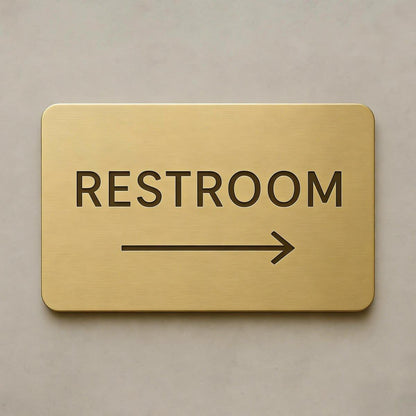 Modern brushed gold toilet direction sign with black text and right arrow displayed on dark marble, grey textured stone and wooden wall backgrounds.
