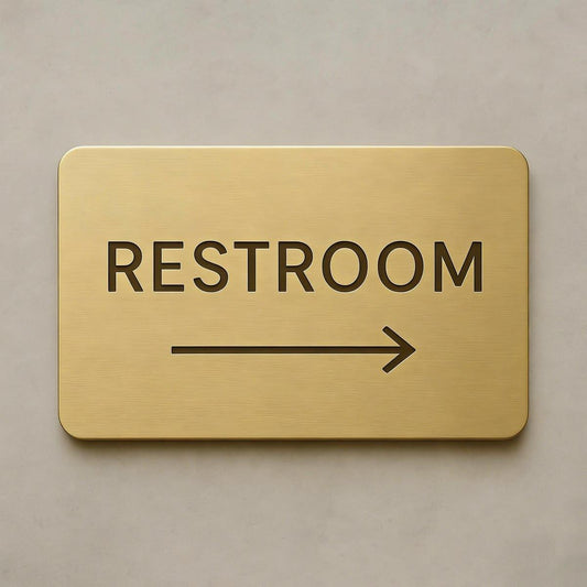 Modern brushed gold toilet direction sign with black text and right arrow displayed on dark marble, grey textured stone and wooden wall backgrounds.
