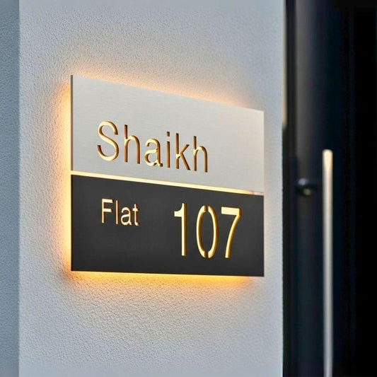 Modern two-tone apartment name plate with brushed gold upper panel, black lower section, and illuminated flat number 107 beside a contemporary entrance.
