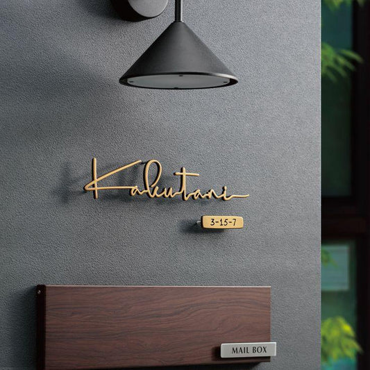 Modern gold cursive house name sign with address number 3-15-7 and a matching wooden mailbox, displayed beside a contemporary home entrance with warm lighting.