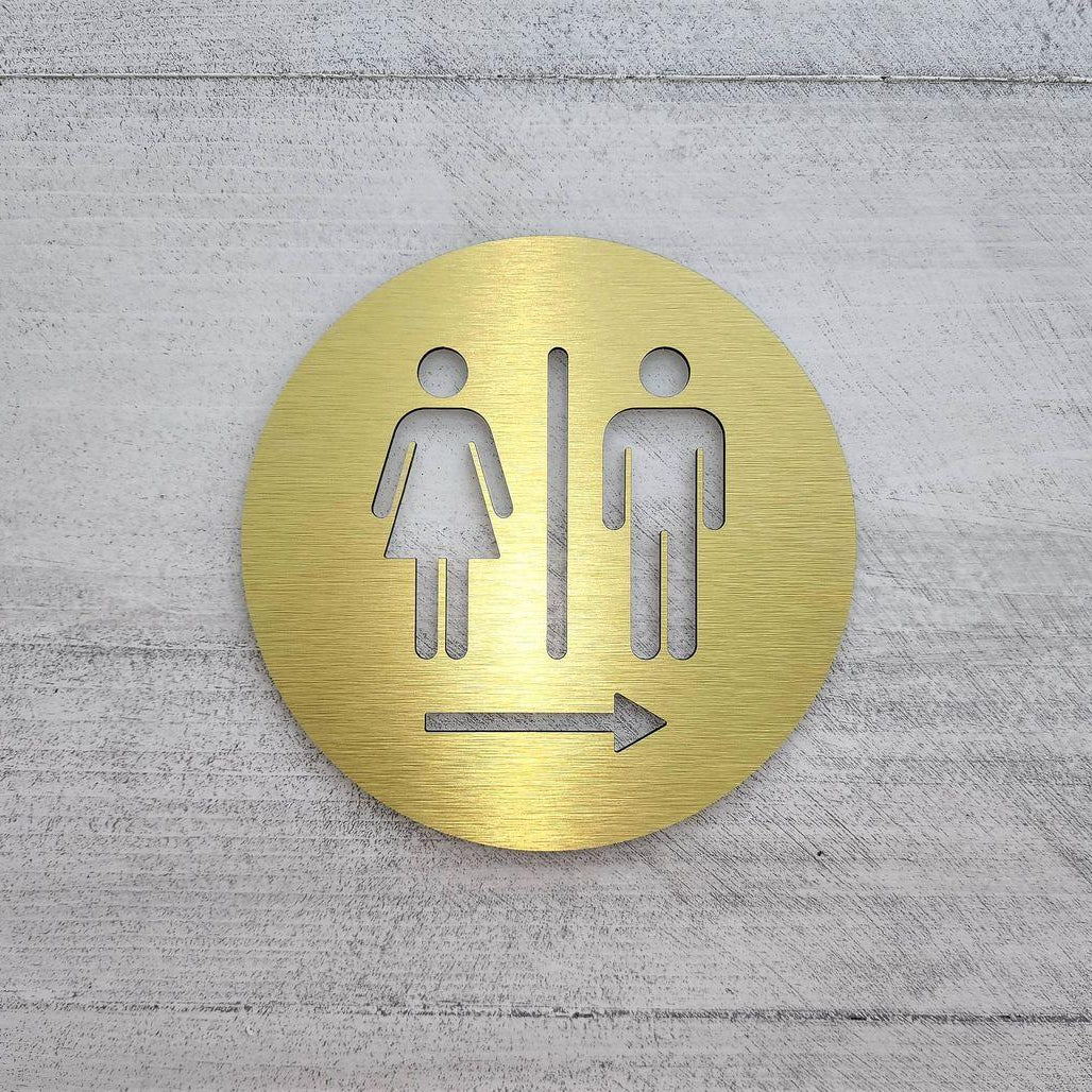 Brushed gold circular toilet signage with engraved-style gender pictograms and directional arrow displayed on wooden panels, dark marble and textured stone walls.