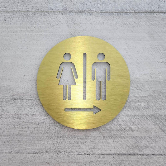 Brushed gold circular toilet signage with engraved-style gender pictograms and directional arrow displayed on wooden panels, dark marble and textured stone walls.