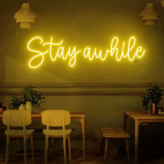 Stay A While Neon Sign | Custom Neon Sign | Neon for Restaurants Neon sign reading 'Stay awhile' in a dimly lit room with tables and chairs.